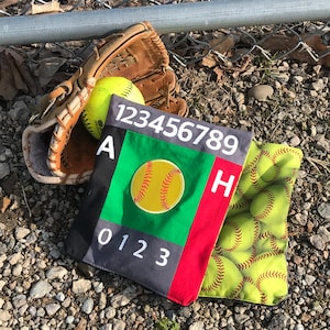 Puede incluir: Una bolsa de tela negra y roja con los números 123456789 y las letras A y H impresas en ella. La bolsa está decorada con una pelota de softball amarilla y tiene un panel verde con los números 0123 impresos en él. Una segunda bolsa con un patrón amarillo y verde de pelotas de softball está encima de la primera bolsa.