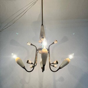 May include: Vintage chandelier with six lights. The fixture has a brass finish with white and cream-colored accents. The light bulbs are visible and lit, casting a warm glow. The design is mid-century modern.