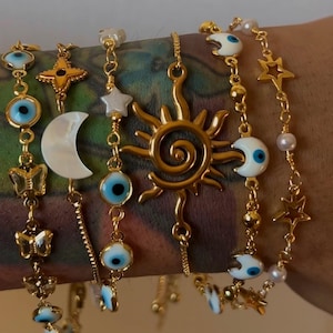 May include: A set of gold chain bracelets with various charms, including evil eyes, stars, moons, butterflies, and a sun. The charms are made of white and blue enamel.