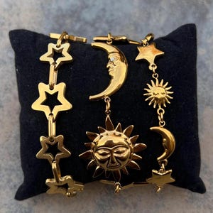 Puede incluir: Pulsera dorada con colgantes celestiales: estrellas, sol y luna. La pulsera se muestra sobre una superficie negra. El sol tiene una cara sonriente, y la luna tiene una cara. Las estrellas son caladas.