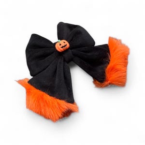 May include: A black fabric bow featuring a small orange pumpkin charm at the centre. The edges of the bow are trimmed with bright orange faux fur, creating a textured contrast against the smooth black material of the main bow structure.