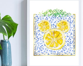 May include: A framed art print featuring three yellow lemon slices arranged on a blue and white floral pattern. The artwork is framed with a gold border and topped with green leaves. The print is displayed on a white surface.