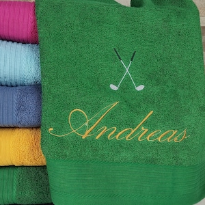 Könnte beinhalten: Ein Stapel Handtücher in verschiedenen Farben, darunter rosa, blau, gelb und grün. Das oberste Handtuch ist grün und hat den Namen "Andreas" in goldener Schrift bestickt, sowie ein Paar gekreuzte Golfschläger.