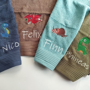 Op de afbeelding: Vier handdoeken met geborduurde dinosaurussen en namen. De handdoeken zijn blauw, bruin, groen en lichtblauw. De namen zijn Nico, Felix, Finn en Phineas.