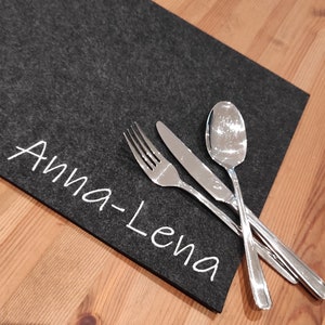 Könnte beinhalten: Ein dunkelgrauer Filz-Tischläufer mit dem Namen "Anna-Lena" in weißer Schrift. Besteck, darunter eine Gabel, ein Messer und ein Löffel, ist auf dem Tischläufer angeordnet, der auf einem Holztisch liegt.