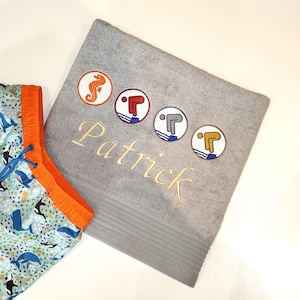 Könnte beinhalten: Ein graues Strandtuch mit gesticktem Seepferdchen, Tauchern und Patrick. Das Handtuch ist in der Hälfte gefaltet und hat ein gestreiftes Muster.