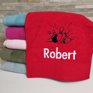 Op de afbeelding: Rode handdoek met een wit geborduurd ontwerp van bowlingpinnen en een bowlingbal. De naam "Robert" is onder de bowlingpinnen geborduurd.