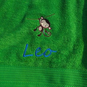 Può includere: Un asciugamano verde brillante con un ricamo di una scimmia e il nome "Leo" in blu. La scimmia è marrone e beige, appesa a una sottile linea. L'asciugamano ha una superficie strutturata.