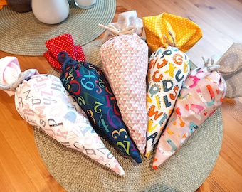 Könnte beinhalten: Eine Sammlung von fünf kegelförmigen Geschenktüten aus Stoff mit verschiedenen Mustern, darunter bunte Buchstaben, Zahlen und geometrische Dreiecke. Jede Tüte ist oben mit einer dicken Kordel zusammengebunden und zeigt Stoffdesigns in Rot, Blau, Rosa, Gelb und Weiß.