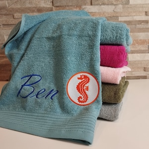 Könnte beinhalten: Ein Stapel blauer, rosa und grüner Handtücher mit dem Namen "Ben" und einem gestickten Seepferdchen auf dem obersten Handtuch.