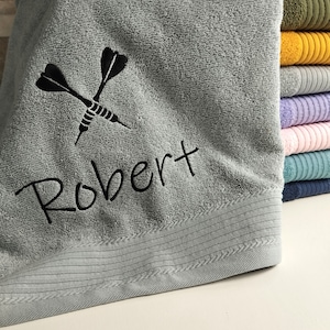 Op de afbeelding: Een lichtgrijze katoenen badhanddoek met een zwart borduursel van twee gekruiste dartpijlen boven de naam Robert in cursief schrift. Een stapel gevouwen handdoeken in diverse kleuren waaronder olijfgroen, geel, grijs, paars, roze en blauw.
