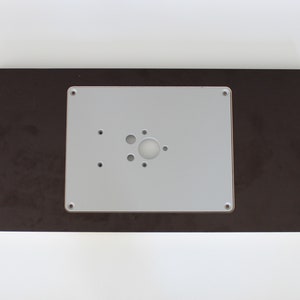 Peut inclure: Une plaque en plastique blanche avec une découpe circulaire et plusieurs petits trous, montée sur une base en bois brun foncé.