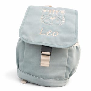 Op de afbeelding: Een lichtblauwe rugzak met een klepsluiting. De klep heeft een geborduurd tijgergezicht en de naam "Leo" in bijpassend lichtbeige garen. De rugzak heeft een zwarte gesp en een beige riem.