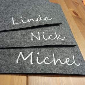 Può includere: Un organizer in feltro grigio con i nomi "Linda", "Nick" e "Michel" ricamati in corsivo bianco. L'organizer ha una forma unica ed è posto su una superficie di legno.