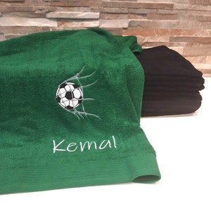 Könnte beinhalten: Ein grünes Handtuch mit einem gestickten Fußball im Netz und dem Namen Kemal in weißem Garn. Ein Stapel gefalteter schwarzer Handtücher befindet sich hinter dem grünen Handtuch, das einen strukturierten Saum am unteren Rand aufweist.