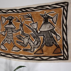 May include: A brown and black textile wall hanging with a geometric design. The design features two figures with large heads and a stylized animal in the center.