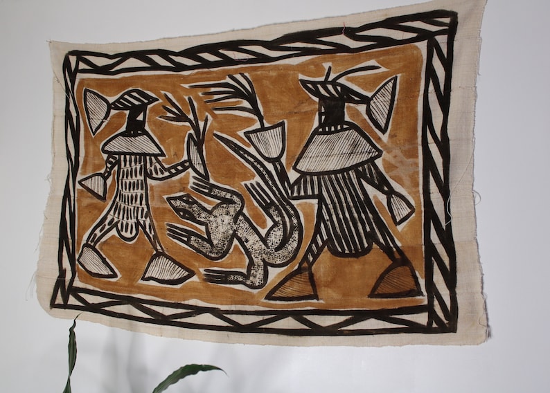 May include: A brown and black textile wall hanging with a geometric design. The design features two figures with large heads and a stylized animal in the center.