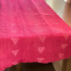 May include: A pink table runner with a geometric pattern of white triangles. The runner has a frayed edge and is made of a woven fabric.