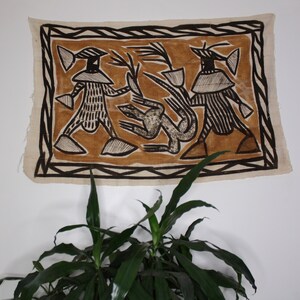 May include: A brown and black textile wall hanging with a geometric design featuring two figures and a stylized animal. The figures are wearing hats and have patterned clothing. The background is a light brown color.