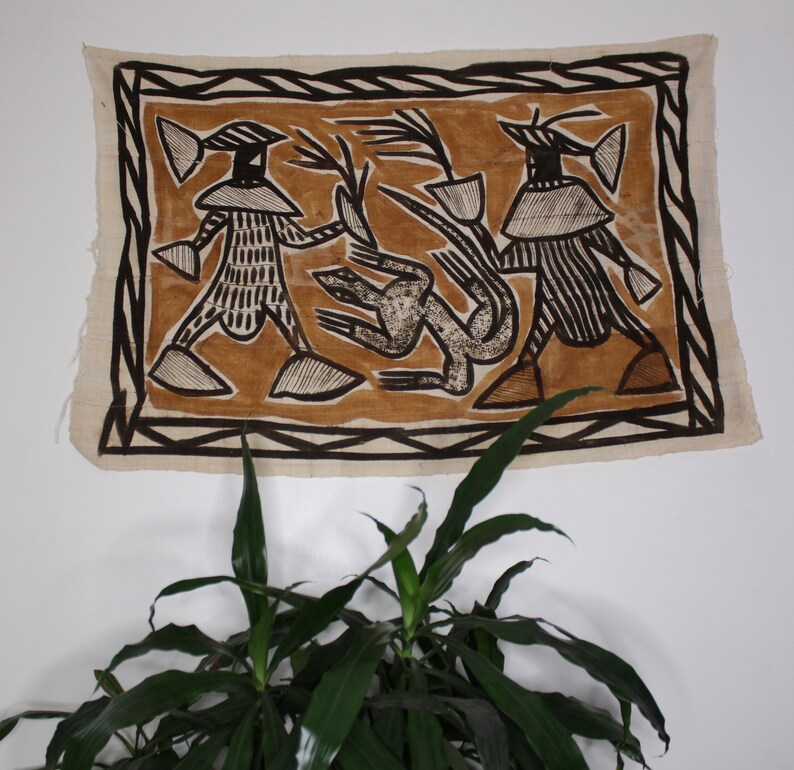 May include: A brown and black textile wall hanging with a geometric design featuring two figures and a stylized animal. The figures are wearing hats and have patterned clothing. The background is a light brown color.