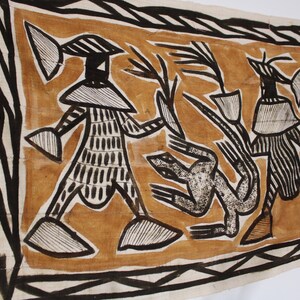 May include: A brown and black textile wall hanging with a geometric design featuring three figures. The figures are stylized and have large heads and bodies. The background is a light brown color.