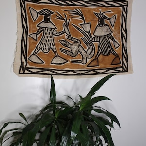 May include: A brown and black textile wall hanging with a geometric design featuring two figures and a bird. The figures are wearing hats and have striped patterns. The background is a light brown color.
