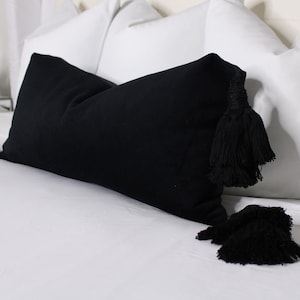 May include: A black pillow with tassels on a white bed. The pillow is made of a soft, textured fabric.