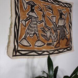 May include: A brown and black textile wall hanging with a geometric design featuring three figures. The figures are stylized and have a tribal aesthetic. The textile is framed with a black border.