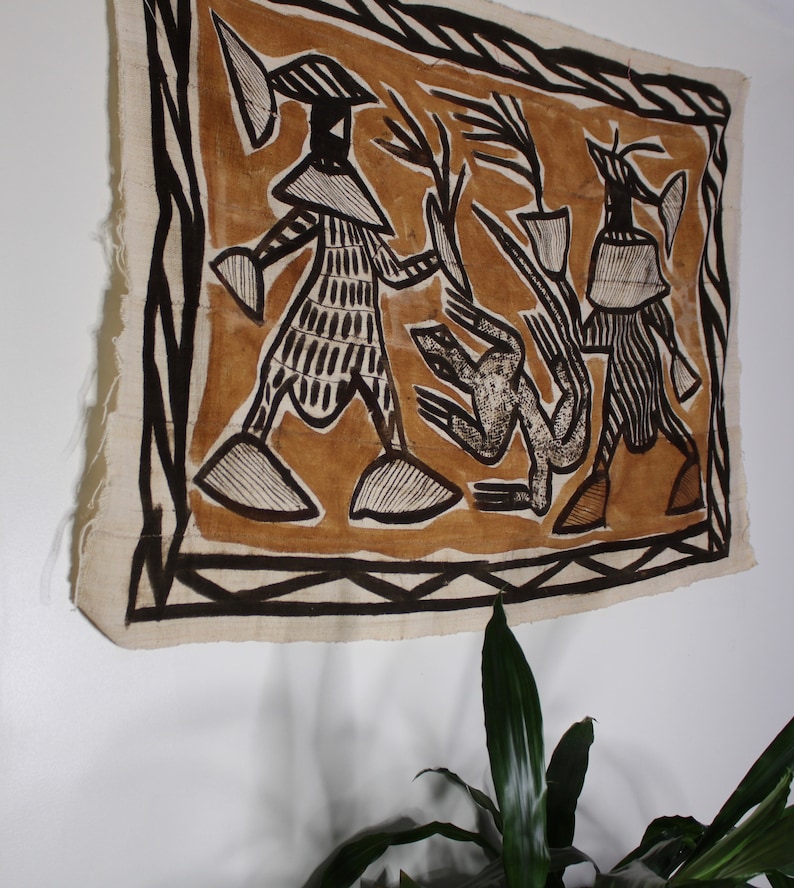 May include: A brown and black textile wall hanging with a geometric design featuring three figures. The figures are stylized and have a tribal aesthetic. The textile is framed with a black border.