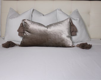 May include: A white bed with a gray velvet pillow with tassels. The pillow is on top of two white pillows.
