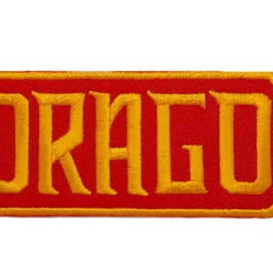 Puede incluir: Parche bordado rojo y dorado con la palabra "DRAGO" en una fuente estilizada.