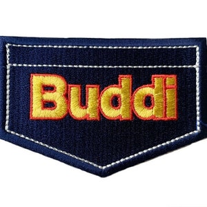 Op de afbeelding: Een blauwe denim patch met witte stiksels in de vorm van een zak. Het woord "Buddi" is geborduurd in geel met een rode omtrek.