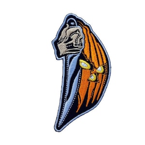 Op de afbeelding: Geborduurde patch met een gestileerd ontwerp met een schedelachtig figuur, oranje en gele accenten en een blauwe en zwarte rand. De patch heeft een unieke vorm en is geschikt voor kleding of accessoires.