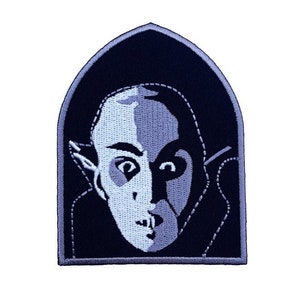 Op de afbeelding: Een geborduurde patch in zwart en wit van een vampier met een puntig oor en een donkere uitdrukking. De patch is boogvormig en heeft een grijze rand.