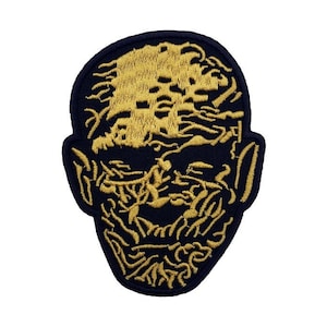 Op de afbeelding: Geborduurde patch met een gestileerd gezicht, een donkerblauwe achtergrond en goudkleurige details. Het ontwerp heeft een textuur, met ingewikkelde steken die de gezichtskenmerken creëren. Geschikt voor kleding of accessoires.