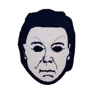 Op de afbeelding: Geborduurde strijkpatch met een wit gezicht met zwarte ogen en een zwarte omtrek. Het gezicht heeft een serieuze uitdrukking en doet denken aan een horrorfilmkarakter.