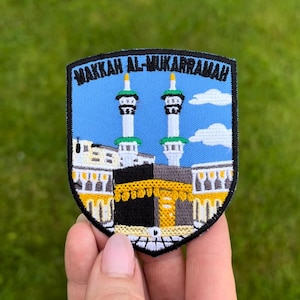 Puede incluir: Parche bordado con la Kaaba en La Meca, Arabia Saudita, con dos minaretes y el texto "MAKKAH AL-MUKARRAMAH" en azul. El parche tiene forma de escudo con un borde negro y está sobre un cielo azul.