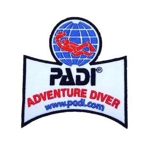 Può includere: Una toppa bianca con un globo blu e una figura rossa che si tuffa. Il testo "PADI" in nero e "ADVENTURE DIVER" in rosso e blu è sotto il globo. Il sito web "www.padi.com" è stampato in blu sotto il testo.