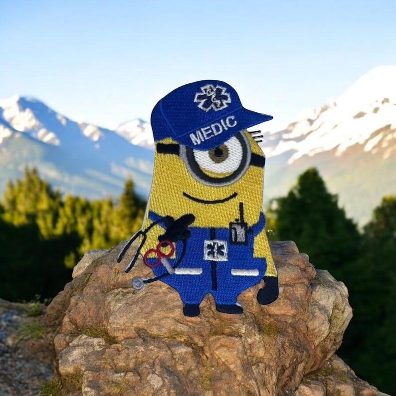 May include: A yellow Minion patch wearing a blue Medic hat and uniform, holding medical tools. The patch has the word "MEDIC" on the hat and a medical symbol. The background shows mountains and trees.