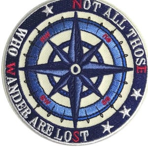 Peut inclure: Un écusson brodé bleu et blanc avec un motif de boussole et le texte "Not all those who wander are lost".