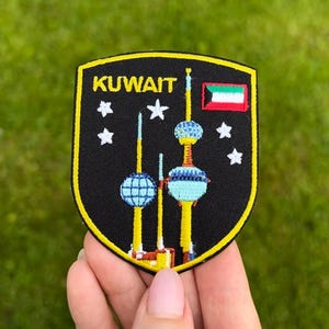 Puede incluir: Parche bordado con la palabra "KUWAIT" en amarillo, sobre un fondo negro con un borde amarillo. El diseño incluye las Torres de Kuwait, estrellas blancas y la bandera kuwaití. El parche se sostiene en una mano.