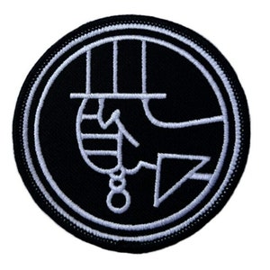 May include: Black and white embroidered patch with a circle design. The design features a stylized hand with a top hat and a rope.