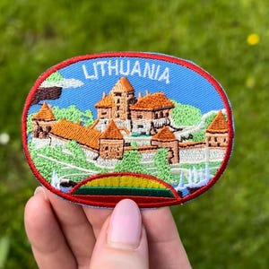 Può includere: Toppa ricamata raffigurante un castello con la scritta "LITHUANIA" in bianco su sfondo blu. La toppa ha un bordo rosso e una striscia verde, gialla e rossa nella parte inferiore, che ricorda una bandiera.