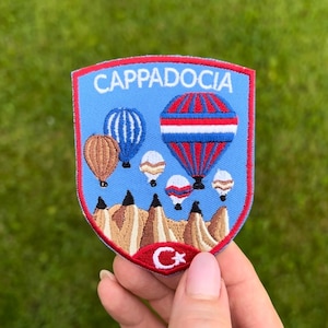 May include: Embroidered patch with the word "CAPPADOCIA" in white on a blue background. The patch shows hot air balloons in red, blue, and brown, above a landscape. A crescent moon and star are at the bottom. Ideal for travel enthusiasts.