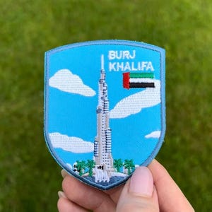 Op de afbeelding: Geborduurde patch met de Burj Khalifa wolkenkrabber in Dubai, Verenigde Arabische Emiraten. De patch heeft een blauwe schildvorm met witte wolken, palmbomen en de tekst "BURJ KHALIFA". De vlag van de VAE is ook inbegrepen.
