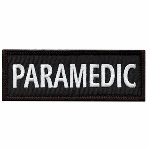 May include: Black rectangular embroidered patch with the word "PARAMEDIC" in white, block letters. The patch has a dark brown border and is likely designed to be sewn onto clothing or gear.