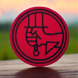 May include: A red embroidered patch with a black outline. The patch features a stylized image of a hand holding a knife.