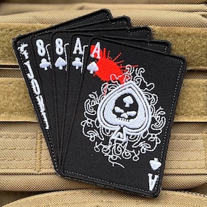 May include: A black patch featuring a white skull and crossbones ace of spades design. The patch has a red blood splatter and is surrounded by a white floral design. The patch is on a brown fabric background.