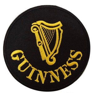 Puede incluir: Un parche redondo negro con texto bordado en oro y un diseño de arpa. La palabra "GUINNESS" está arqueada en la parte inferior. El arpa está centrada encima del texto.