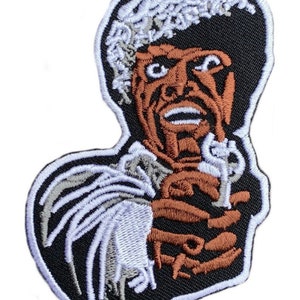 Puede incluir: Un parche bordado para planchar que presenta un personaje de dibujos animados con una afro blanca, piel marrón y una camisa blanca. El personaje sostiene una pistola en su mano derecha y tiene una expresión de sorpresa en su rostro.